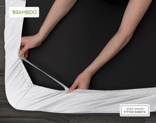 Bottom view of our Bamboo Cotton Fitted Sheet in White to show two hands pulling the Snug Fit elastics holding the corner in place over a mattress.