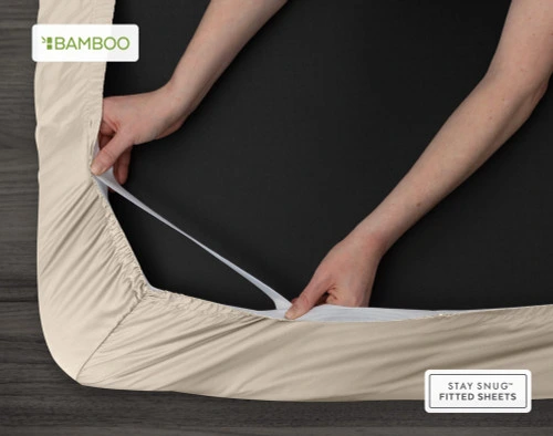 Bottom view of our Bamboo Cotton Fitted Sheet in Driftwood Beige to show two hands pulling the Snug Fit elastics holding the corner in place over a mattress.