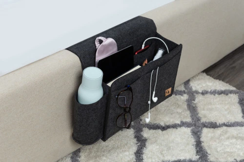 Side view of our Bedside Felt Pocket in Dark Grey with living room essentials like a remote, tablet, and water bottle.