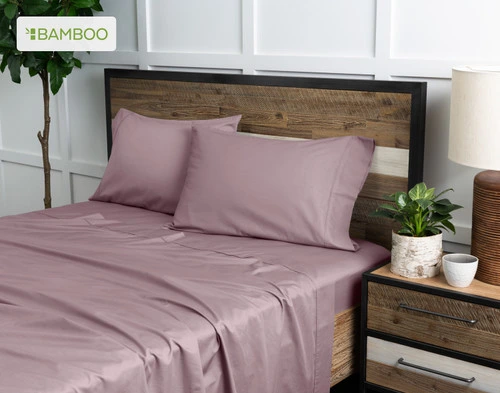 Angled view of our Bamboo Cotton Sheet Set in Orchid Purple dressed over a wooden bed in a white plant-filled bedroom.