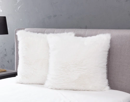 Angled view of two Polar Fur Euro Shams in White resting against a headboard on an empty bed in a light grey bedroom.