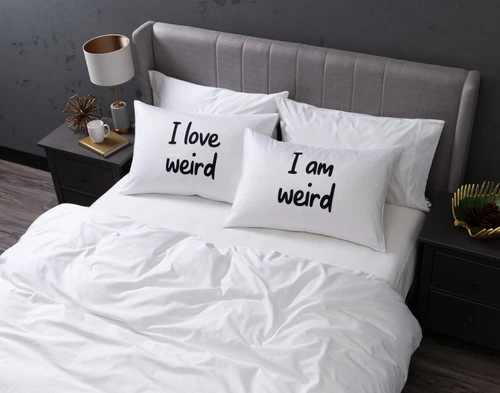 Angled view of our I Love Weird Pillow Talk Pillowcases sitting on a white bed in a dark grey bedroom.