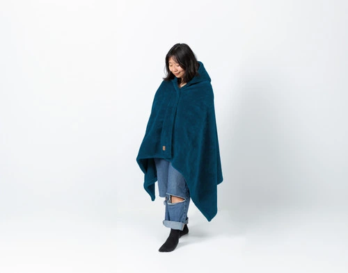 Angled view of a young woman looking downwards while wearing our Hooded Cape Plush Wearable Throw in Teal.