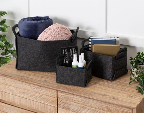 Angled view of our three Collapsible Felt Basket sizes holding various books, household wares, and cushions on top of a dresser.