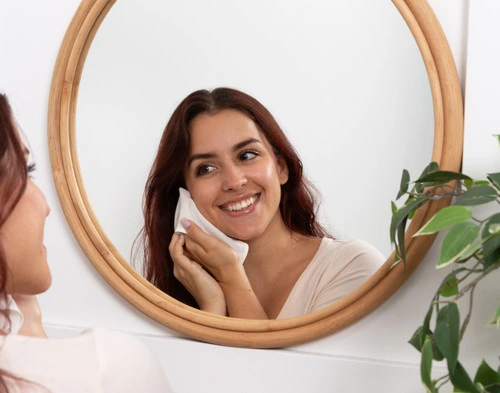 Woman cleansing her face with our Silkglow Face Cloth while smiling in a mirror.