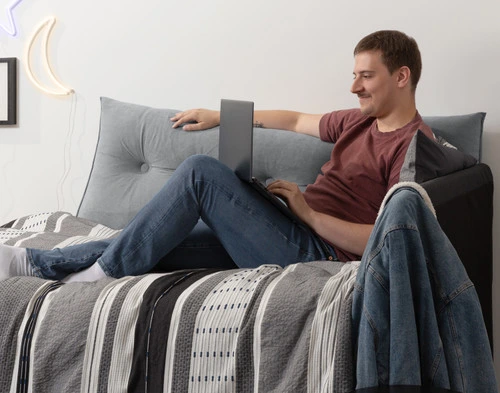 Angled view of a twin bed being used as a couch with our Grey Wedge Pillow against the wall, with a young male student lounging on it while browsing his laptop.