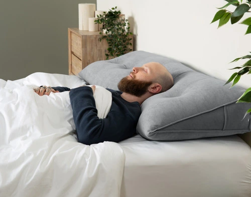 Side view of a white bed with our Grey Wedge Pillow laying on its face, providing angled support for a sleeping man laying on his back.