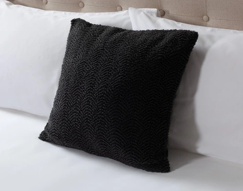 Angled view of our Ripple Boucle Square Cushion in Soot leaning against two white pillows on an undressed bed.