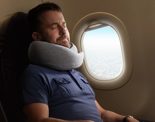 Bearded man sleeping on an airplane wearing our Neck-Hug Travel Pillow next to an open window.