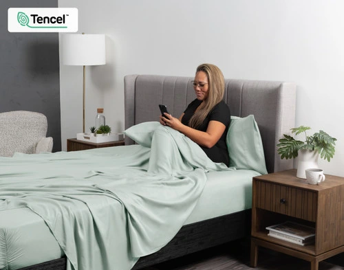 Angled view of the BeechBliss Tencel Modal Sheet Set in light green Waterfall dressing a bed with a woman on her phone resting in it.