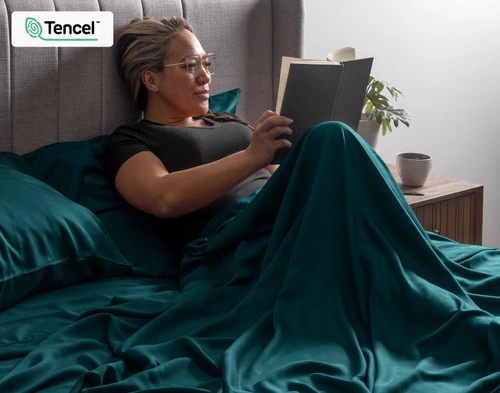 Woman sitting upright in a bed reading her book curled up in the flat sheet of our BeechBliss TENCEL™ Modal Sheet Set in Jungle.