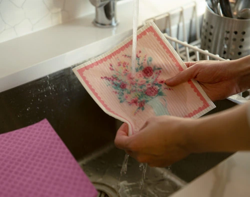 Angled view of our Pitcher Bouquet Swedish Dishcloth under a sink being sprayed with water before cleaning.