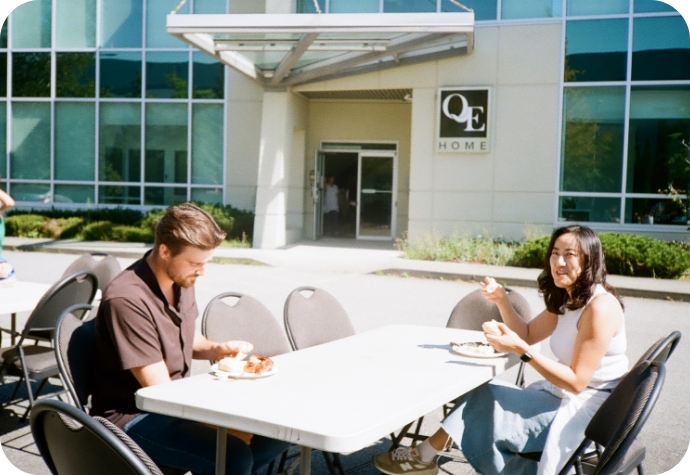 Our Chief Operations Officer and Sustainability Manager sit at a table enjoying their lunch, with the QE Home logo visible on the office exterior in the background.