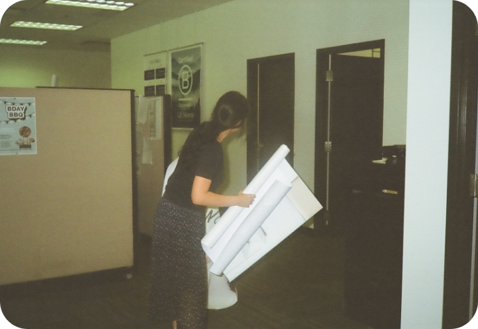 A QE Home graphic designer manages multiple print signs in the office, with an existing store B Corp poster visible in the background.