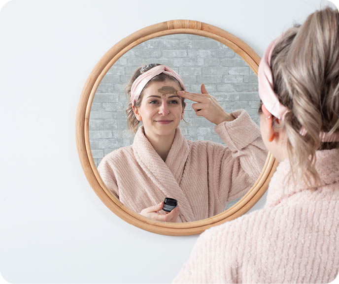 A person applies skincare to their face in front of a round mirror, wearing a pink plush robe and silk headband.