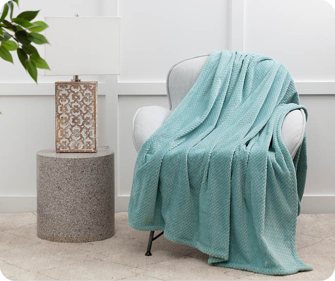 Our Chevron Plush Throw in Aqua shown draped over an armchair in a room next to a side table and lamp.