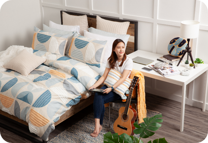 A woman with a tablet sits by a bed dressed in our Nora Duvet Cover and Pillow Shams in a modern, well-decorated dorm room.