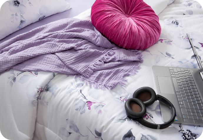 A bed with our floral Talia Comforter Set, our Muslin Gauze Throw in Lilac, our pink Sulani Round Cushion, a laptop, and headphones.