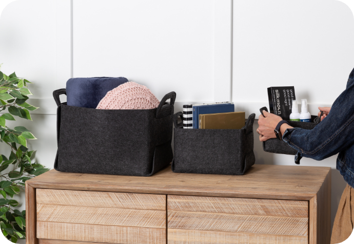 A person organizes items in two our Collapsible Felt Baskets on a wooden dresser beside a green plant.