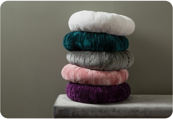 A stack of our Pleated Velvet Round Cushions placed on a velvet bench against a grey wall.