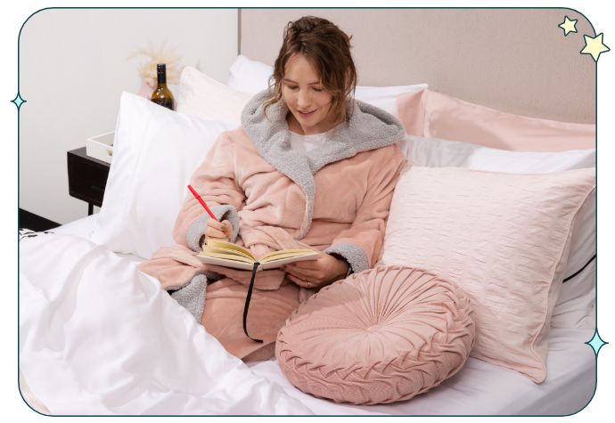 Blonde woman in a pink bathrobe writing happily in her journal from bed.