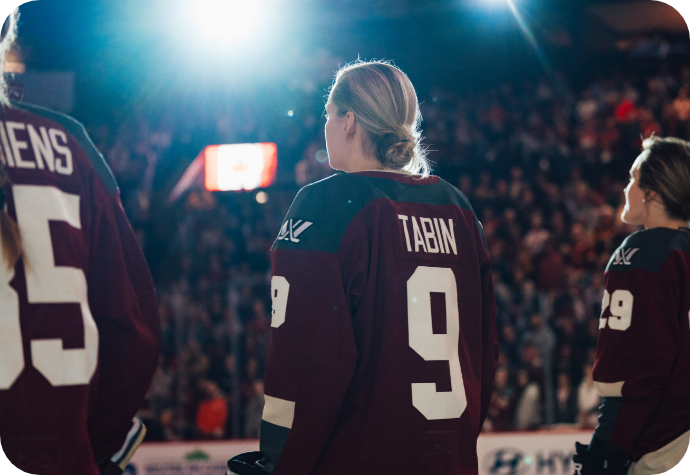 Hockey player Kati Tabin wearing the official PWHL Montreal maroon jersey with the name "Tabin" facing away from the camera, with teammates. Bright arena lights shine above, and a blurred crowd is visible in the background.