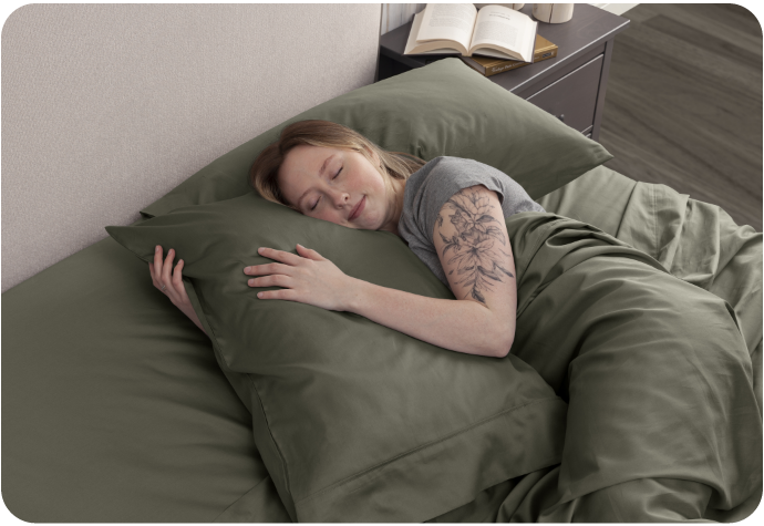 Angled view of a woman sleeping in a green bed while wrapping her arms around a pillow.